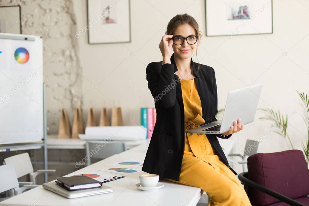 Young beautiful woman in eyeglasses and black jacket sitting on desk happily looking in camera with laptop in hands and cup of coffee near at work in modern office