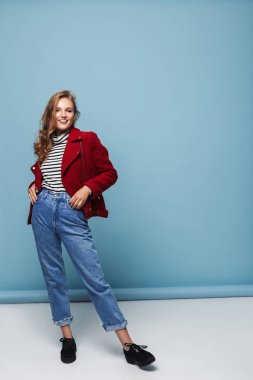 Kırmızı ceket ve kot sevinçle kamera tutuşup ceplerinde mavi arka plan üzerinde seyir dalgalı saçlı Genç mutlu kadın