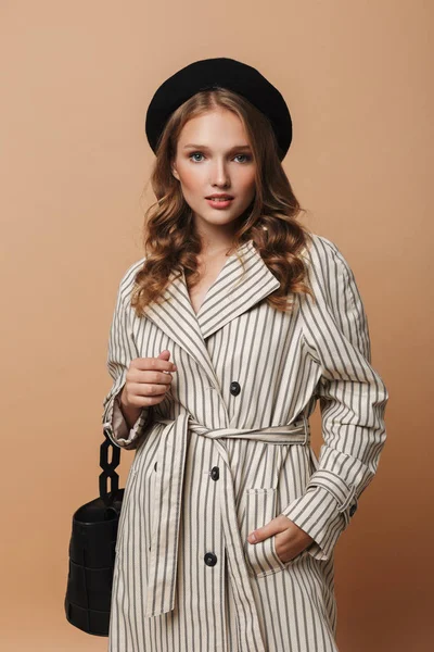 Young pretty woman with wavy hair in striped coat and black hat with handbag dreamily looking in camera over beige background