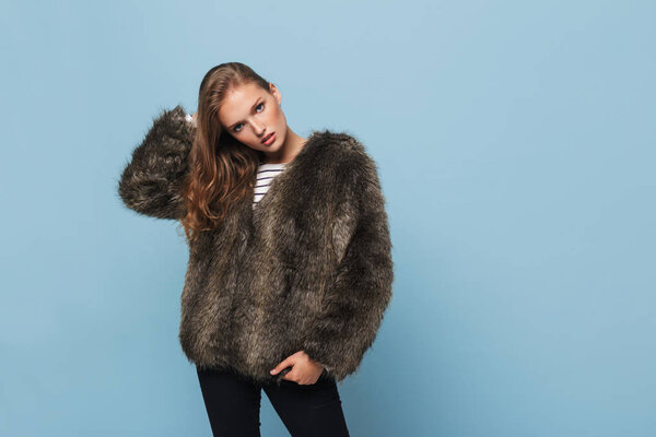 Молодая красивая леди с волнистыми волосами в меховой куртке задумчиво глядя в камеру на голубом фоне
