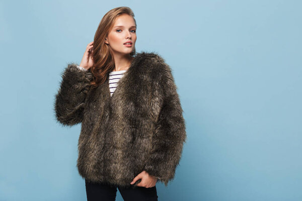 Красивая девушка с волнистыми волосами в меховой куртке мечтательно смотрит в камеру на синем фоне
