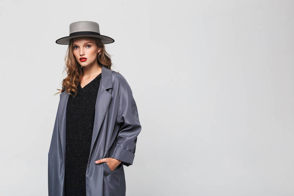 Молодая красивая женщина с волнистыми волосами в шляпе и плащом, держась за руки в карманах мечтательно глядя в камеру на сером фоне
