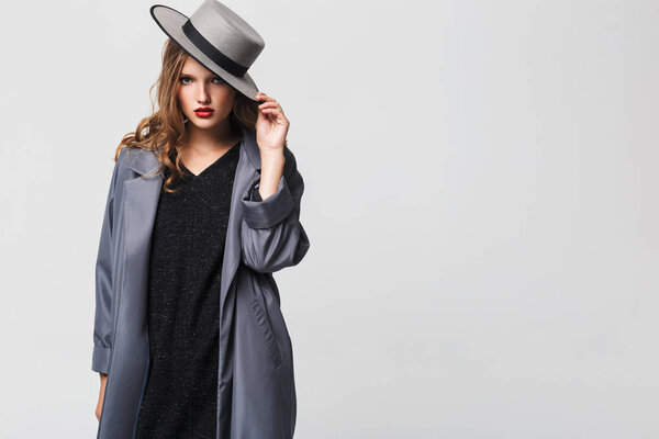 Молодая красивая женщина с волнистыми волосами в шляпе и плаще мечтательно глядя в камеру на сером фоне
