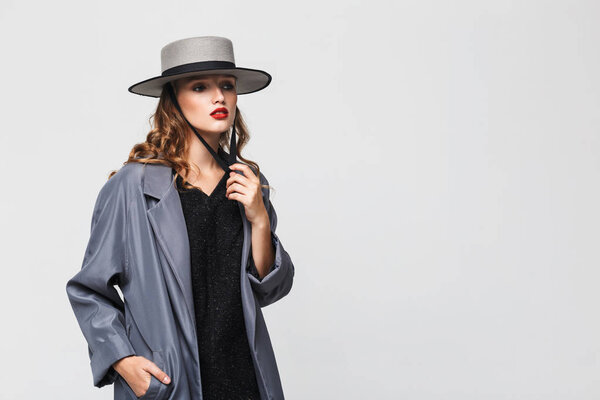 Молодая привлекательная задумчивая женщина с волнистыми волосами в современной шляпе и плащом мечтательно глядя в сторону на сером фоне
