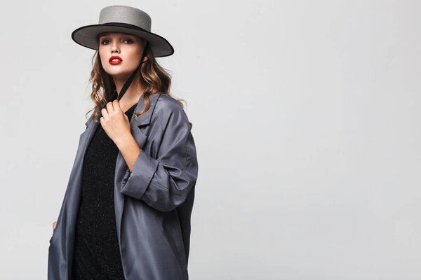 Красивая задумчивая девушка с волнистыми волосами в шляпе и плащом мечтательно глядя в камеру на сером фоне
