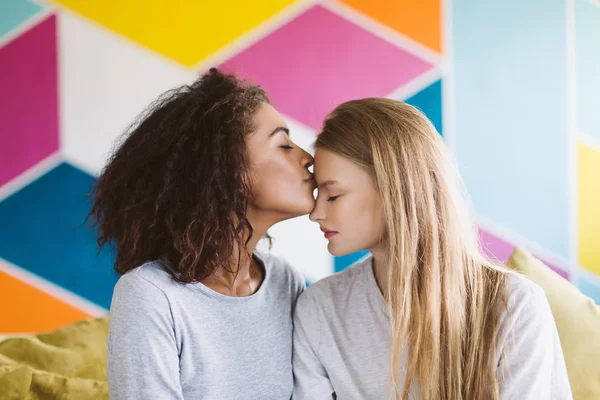 Beautiful african american woman with dark curly hair dreamily kissing forehead of pretty woman with blond hair with colorful wall on background