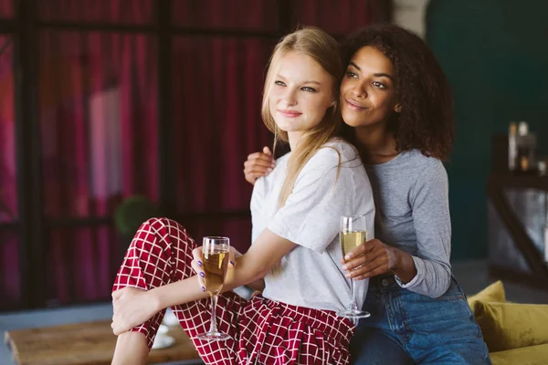 Beautiful african american woman with dark curly hair and pretty woman with blond hair leaning on each other with glasses of champagne in hands dreamily looking aside at home