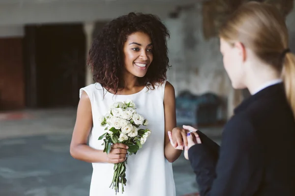 年轻有吸引力的非洲裔美国妇女与深色卷发在白色的礼服举行小花束鲜花在手快乐地花时间在婚礼上 — 图库照片