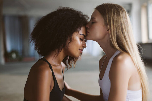 Close up young woman with blond hair thoughtfully kissing in forehead smiling african american woman with dark curly hair spending time together at home