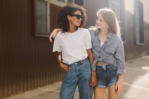 Young african american woman with dark curly hair in sunglasses and T-shirt and pretty woman with blond hair in shirt dreamily looking at each other spending time together