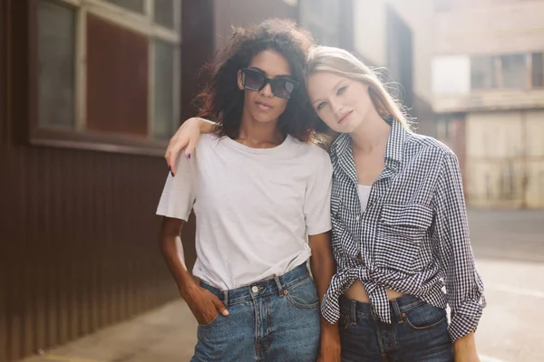 Young african american woman with dark curly hair in sunglasses and T-shirt and pretty woman with blond hair in shirt dreamily looking in camera spending time together