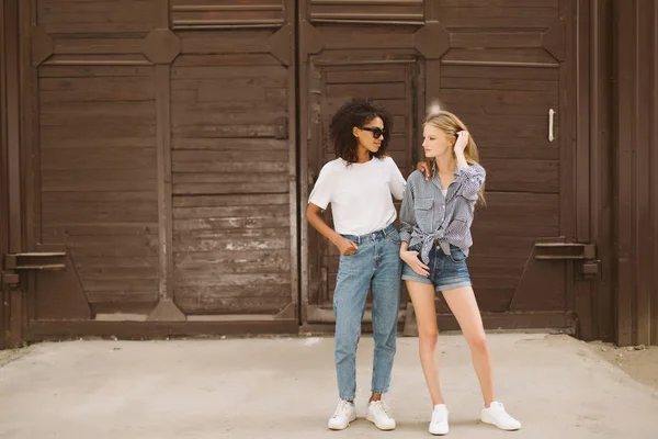 Young african american woman with dark curly hair in sunglasses and T-shirt and nice woman with blond hair in shirt thoughtfully looking aside spending time together outdoor