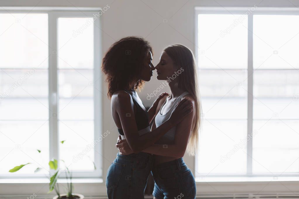 Young beautiful woman with blond hair and pretty african american woman with dark curly hair dreamily hugging each other at home with windows on background