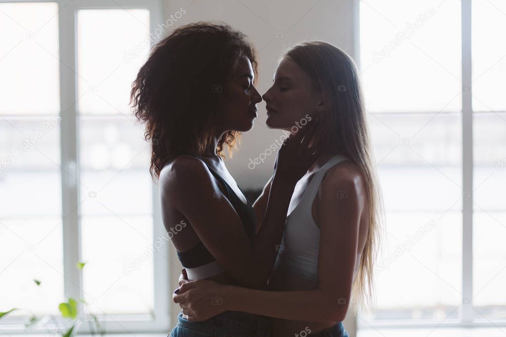 Young attractive woman with blond hair and african american woman with dark curly hair dreamily hugging each other spending time together at home with windows on background