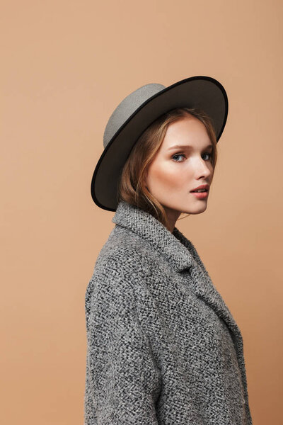 Портрет молодой красивой женщины с волнистыми волосами в серой шляпе и пальто, мечтательно смотрящей в камеру на бежевом фоне
