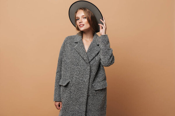 Молодая красивая привлекательная женщина с волнистыми волосами в серой шляпе и пальто мечтательно глядя в камеру на бежевом фоне
