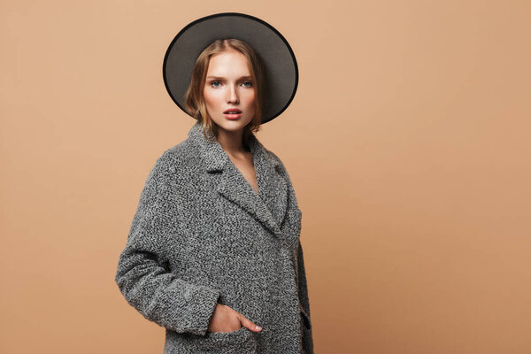 Молодая красивая леди с волнистыми волосами в серой шляпе и пальто держа руку в кармане мечтательно глядя в камеру на бежевом фоне
