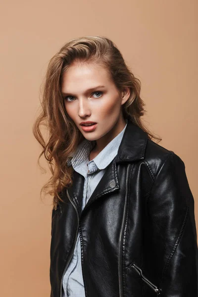 Young beautiful woman with wavy hair in black leather jacket and shirt thoughtfully looking in camera over beige background