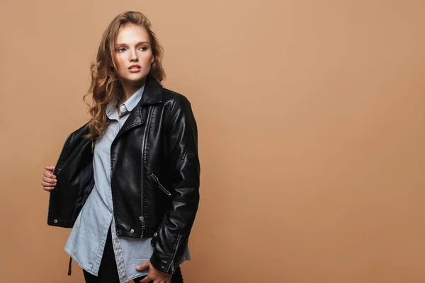 Young attractive woman with wavy hair in black leather jacket and shirt thoughtfully looking aside over beige background