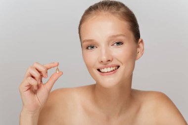 Portrait of beautiful smiling girl without makeup holding little capsule pill in hand happily looking in camera over gray background clipart