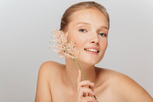 Портрет молодой улыбающейся девушки без макияжа, держащей красивую ветку возле лица, мечтательно смотрящей в камеру на сером фоне
