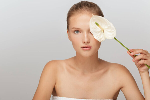 Портрет привлекательной женщины без макияжа, закрывающий глаза белым цветком каллы, задумчиво смотрящей в камеру на сером фоне
