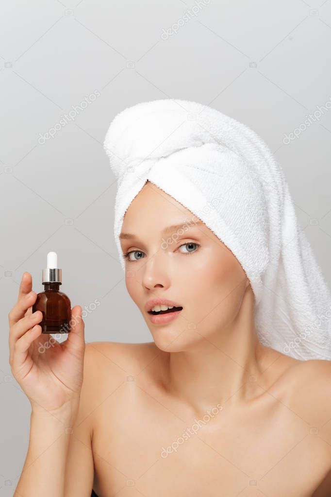 Portrait of young lady without makeup with white towel on head holding liquid bottle in hand amazedly looking in camera over gray background