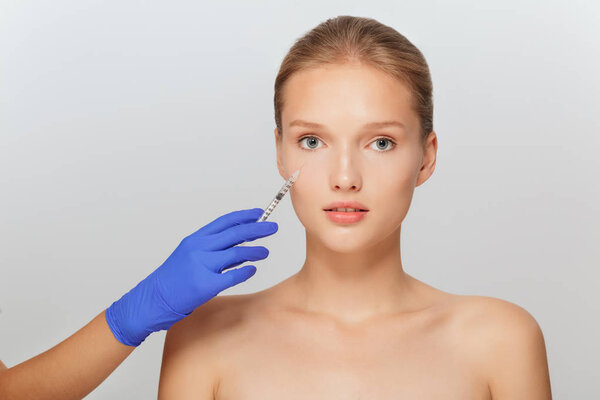 Портрет молодой привлекательной женщины, получающей инъекции красоты в область глаз со шприцем вдумчиво смотрящей в камеру на сером фоне
