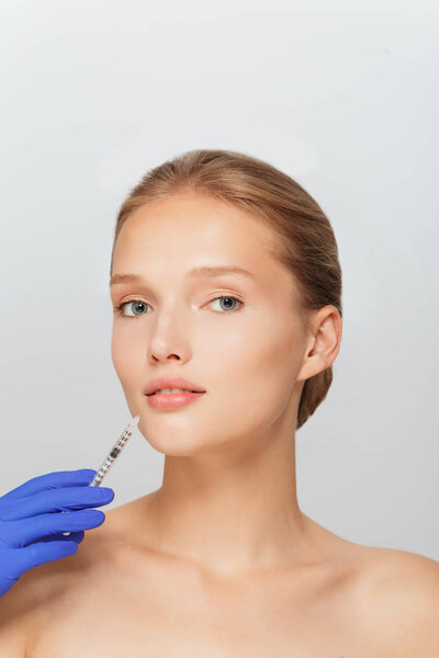 Портрет молодой привлекательной женщины, получающей инъекцию красоты в область рта со шприцем, мечтательно смотрящей в камеру на сером фоне
