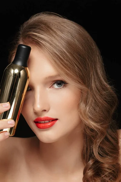 Close up beautiful girl with wavy hair and red lips covering eye with cosmetic bottle dreamily looking in camera over black background