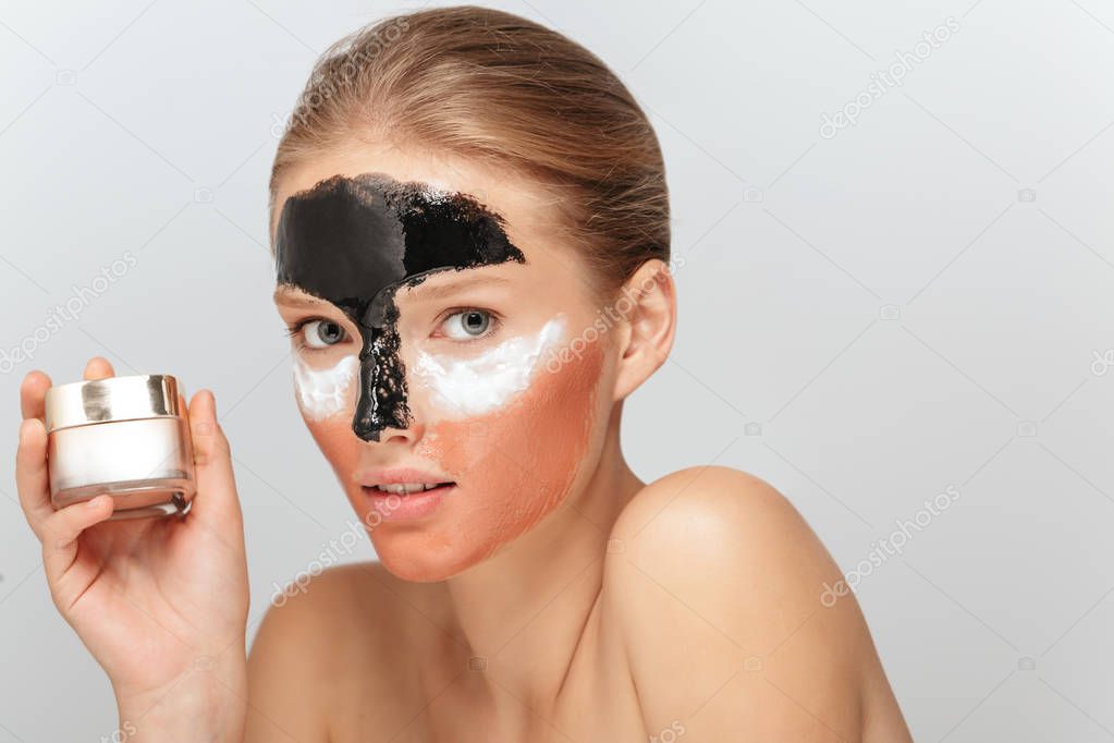 Portrait of young attractive woman with different cosmetic masks on face holding beauty cream in hand thoughtfully looking in camera over gray background