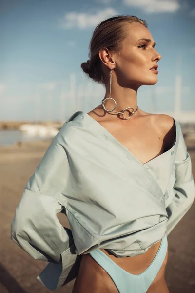 Young beautiful lady in shirt and swimsuit wearing modern silver earrings and neck jewelry dreamily looking aside spending time on beach