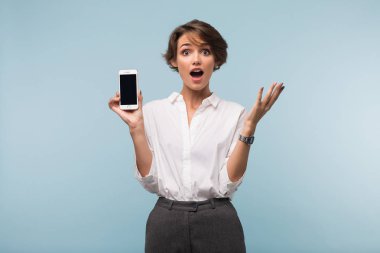 Bayan beyaz gömlek cep telefonu elinde amazedly kamera üzerinde mavi arka plan açık ağız görünmenin tutarak siyah kısa saçlı Genç hayret