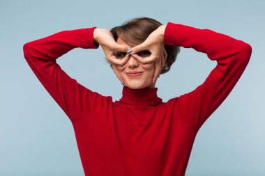 Kırmızı kazaklı sevinçle yapıyor gözlük hareketi el ile mavi arka plan üzerinde siyah kısa saçlı Genç güzel kadın 