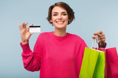 Pembe kazaklı holding renkli alışveriş torbaları ve kredi kartı eller mutlu seyir arıyor kamera mavi arka plan üzerinde siyah kısa saçlı Genç neşeli kadın
