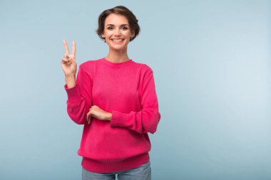 Pembe kazaklı up sevinçle kamera arıyor mavi arka plan üzerinde iki parmak gösterilen siyah kısa saçlı Genç neşeli kadın