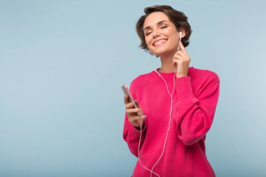 Pembe kazaklı mavi arka plan üzerinde kulaklık el mutlu bir şekilde dinleme müzik cep telefonu tutan güzel rüya kız