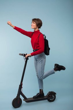 Süper kahraman poz elektrikli scooter mutlu bir kenara seyir üzerinde mavi arka plan üzerinde siyah sırt çantası ayakta neşeli Bayan kırmızı kazak ve kot pantolon