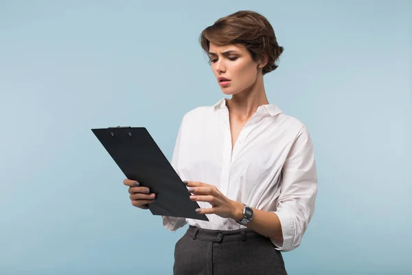 Young serious business woman with dark short hair in white shirt sadly looking on folder with documents over blue background