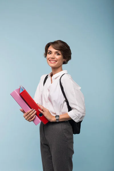 Beautiful smiling girl in white shirt and pants with backpack happily looking in camera holding colorful folders in hands over blue background