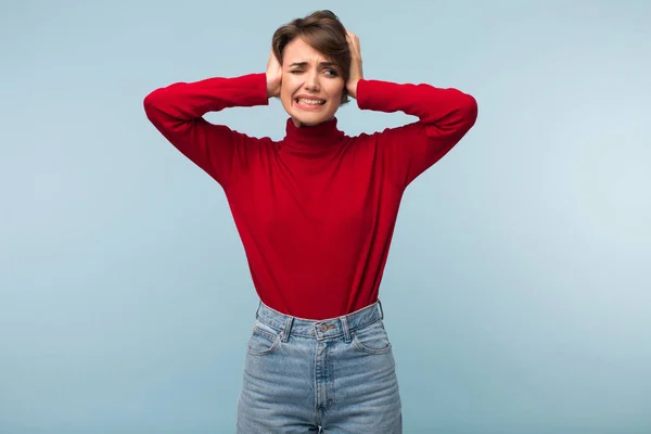 赤いセーターとジーンズで耳を閉じる悲しいことに若いイライラ疲れている女性の手に青い背景に — ストック写真