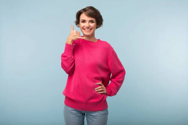 ピンクのセーターとジーンズの青い背景の上カメラで見て楽しく電話ジェスチャーを示す黒い短い髪の若い魅力的な女性 — ストック写真