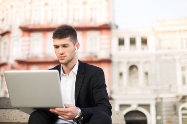 Young serious businessman in classic black suit and white shirt with wireless earphones thoughtfully working on laptop outdoor