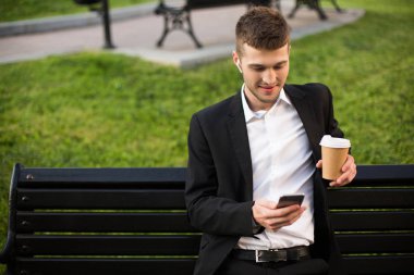 Klasik siyah ceketli, beyaz gömlekli, kablosuz kulaklıklı ve kahveli genç yakışıklı adam bankta otururken mutlu bir şekilde cep telefonunu açık havada kullanıyor.