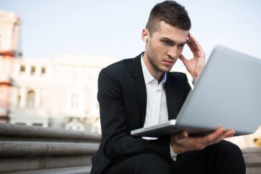 Young serious businessman in classic black jacket and white shirt with wireless earphones thoughtfully working on laptop spending time outdoor