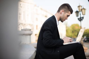Klasik siyah takım elbiseli, kablosuz kulaklıklı genç ve yakışıklı bir iş adamı laptopuyla dışarıda özenle çalışıyor.