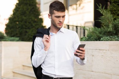 Kablosuz kulaklık siyah ceket düşünceli cep telefonu açık kullanarak omzuna holding ile beyaz gömlekli yakışıklı delikanlı