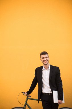 Klasik siyah takım elbise ve beyaz gömlek ile kablosuz kulaklık dizüstü sevinçle kamera ile bisiklet turuncu arka plan üzerinde arıyorum elinde tutan genç neşeli adam