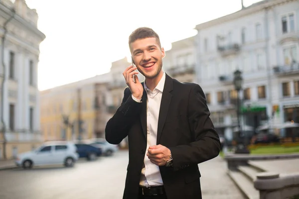 Young handsome smiling man in black jacket and white shirt happily looking aside talking on cellphone with beautiful city view on fuzzy background