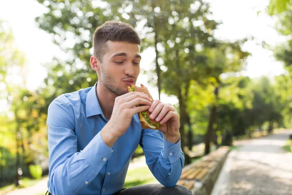 Joven Hombre Atractivo Camisa Azul Sentado Banco Comiendo Alegremente Sándwich — Foto de Stock